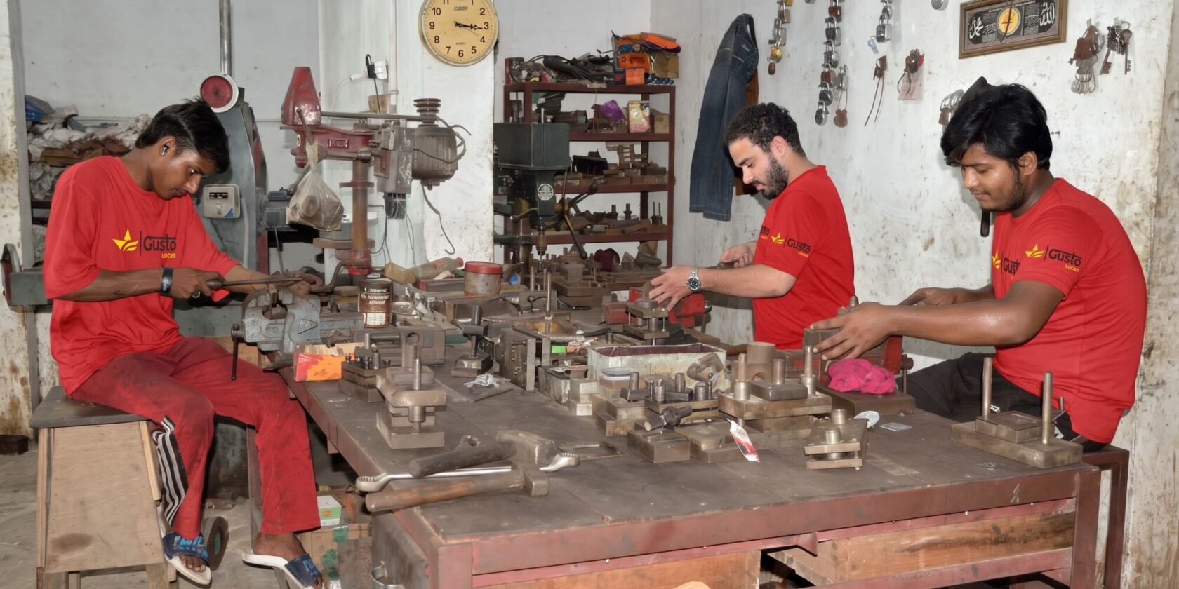 Three workers in Gusto Locks uniform machining and fitting padlock parts at a bench